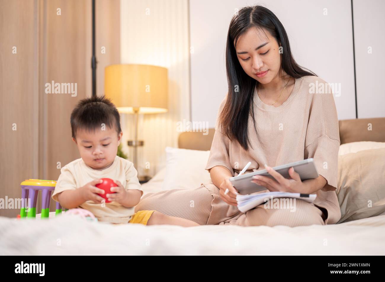 Eine schöne asiatische Mutter arbeitet an ihrem digitalen Tablet, während ihr Bay Boy Spielzeug im Bett spielt. Mamas Lebenskonzept Stockfoto