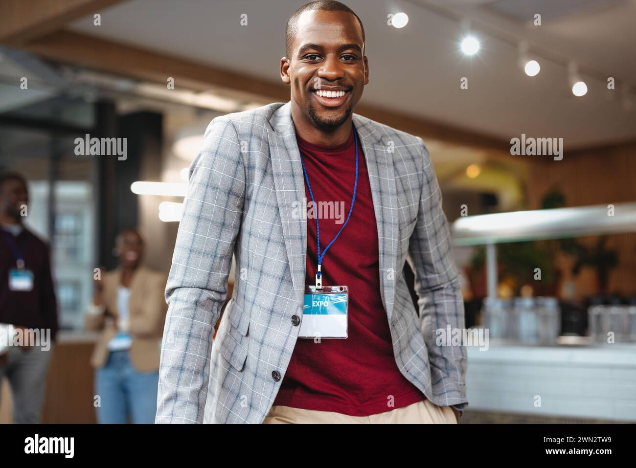 Ein erfolgreicher Geschäftsmann mit einem Abzeichen lächelt während eines Networking-Events. Die Büroumgebung mit Kollegen im Hintergrund spiegelt ein professionelles und wider Stockfoto