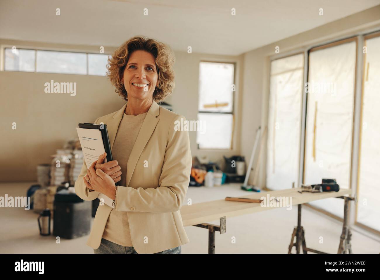 Glückliche Hausbesitzerin steht in ihrer Küche und hält Papiere für eine Hausrenovierung. Mit einem Lächeln schaut sie selbstbewusst in die Kamera und zeigt h Stockfoto