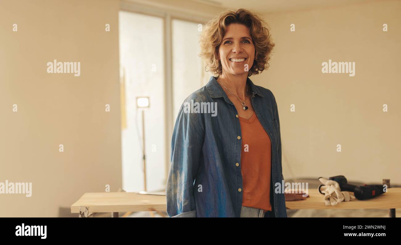 Porträt einer glücklichen Frau, die in ihrem Haus steht und in die Kamera lächelt, während sie sich mit Tischlerei beschäftigt. Reife Frau renoviert ihr Haus, um einen zu erschaffen Stockfoto