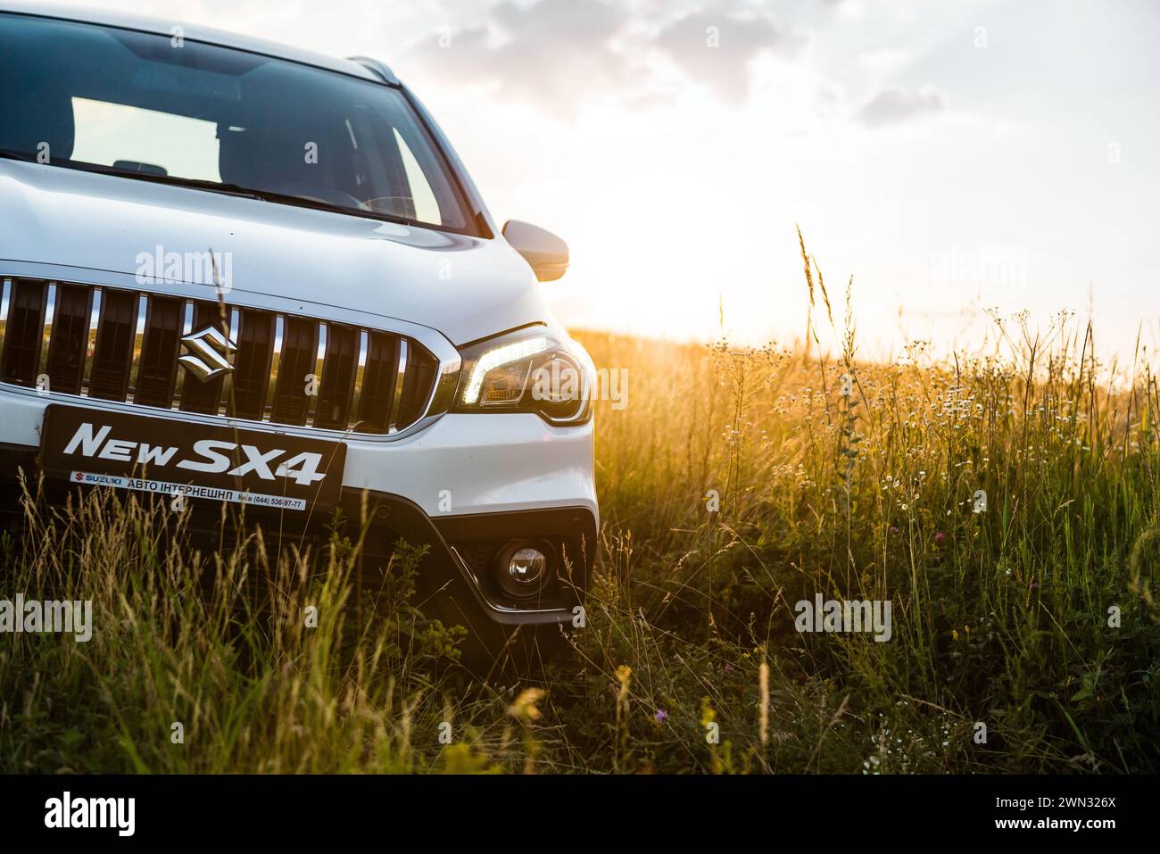 Weißer Suzuki SX4 in einem hügeligen Feld bei Sonnenuntergang. Nahaufnahme der Vorderseite der kompakten Überkreuzung auf einer Wiese - Wochenende weg von der Stadt Stockfoto