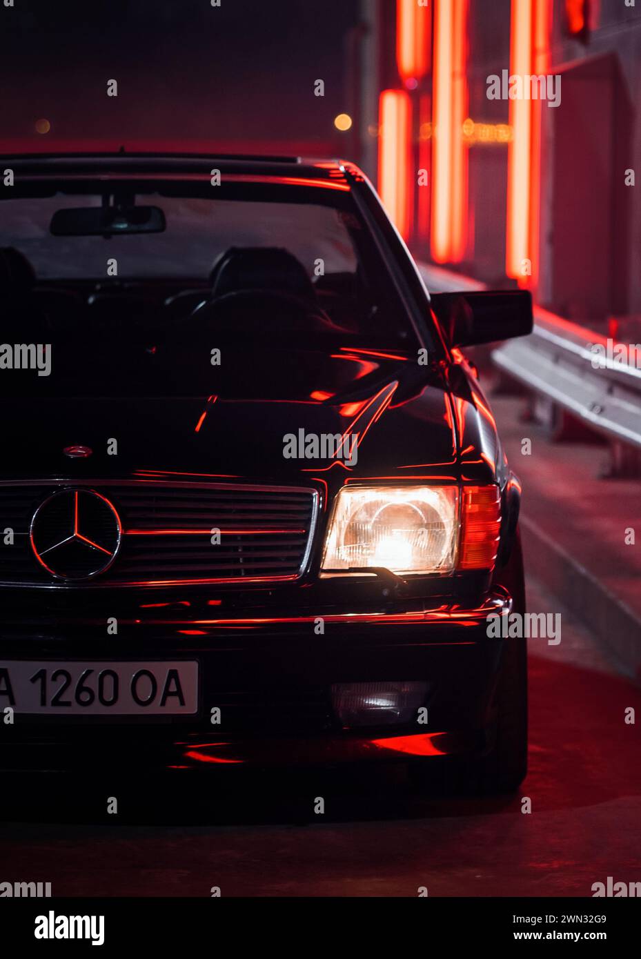 Klassischer Mercedes-Benz W126 auf einer Straße mit rot leuchtenden Lichtern. Vorderansicht eines Luxuswagens aus den 1980er Jahren bei Nacht - Scheinwerfer eingeschaltet Stockfoto