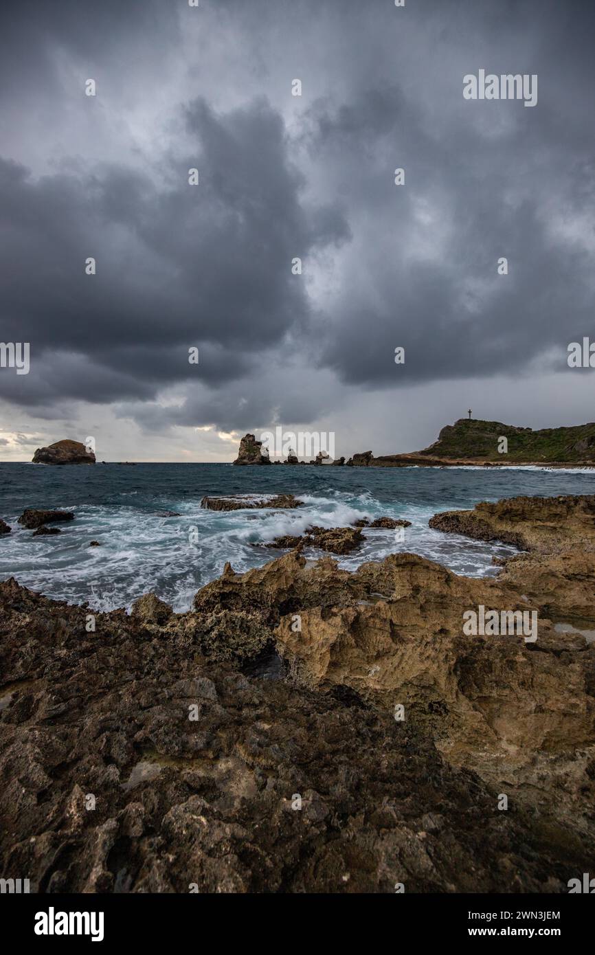 Eine felsige Küste mit einer spitzen Felsformation erstreckt sich in einer Bucht ins Meer, starke Wellen bei einem regnerischen Sonnenaufgang schaffen eine dramatische Atmosphäre. Punkt Stockfoto