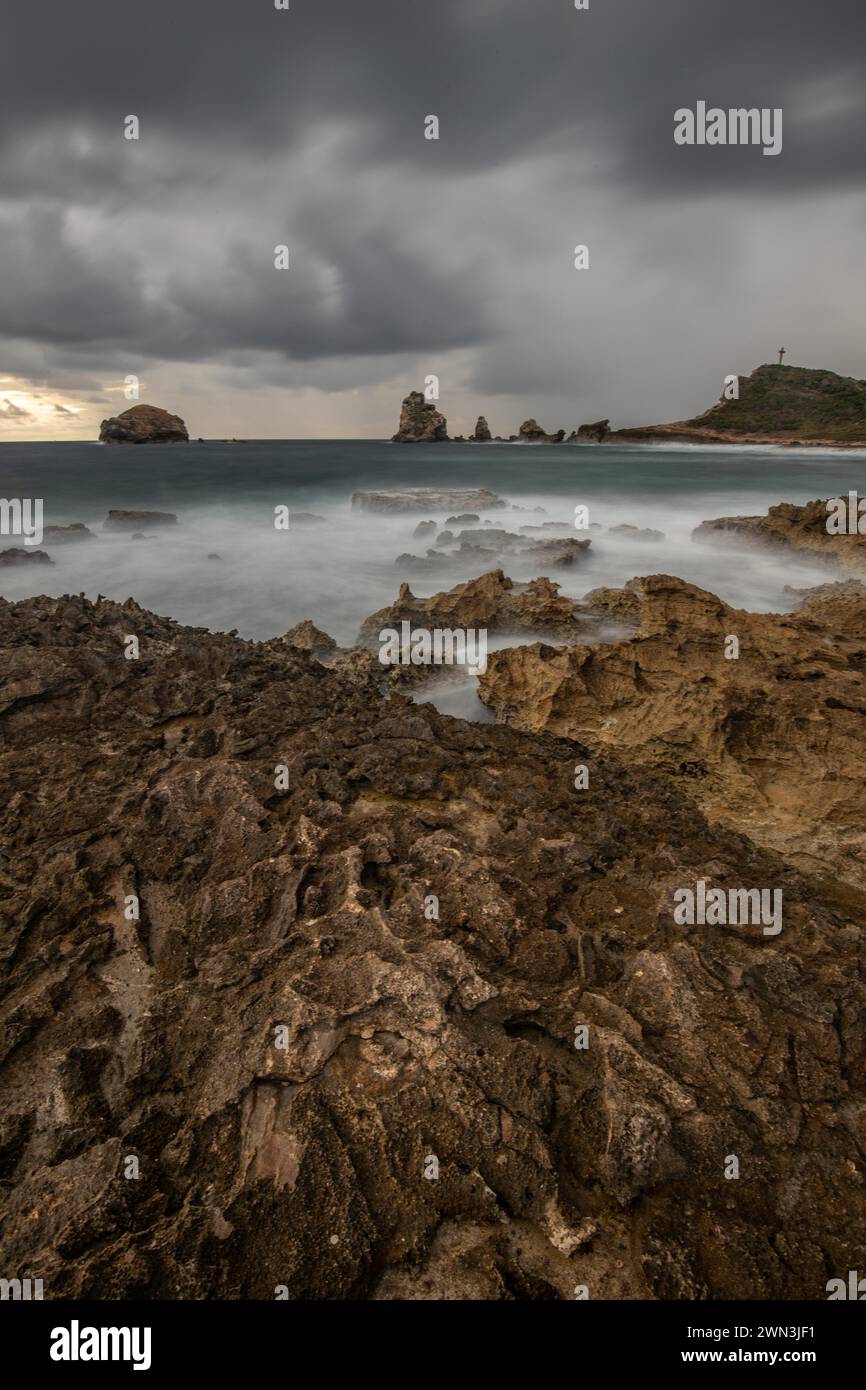 Eine felsige Küste mit einer spitzen Felsformation erstreckt sich in einer Bucht ins Meer, starke Wellen bei einem regnerischen Sonnenaufgang sorgen für eine dramatische Stimmung auf lange Sicht Stockfoto