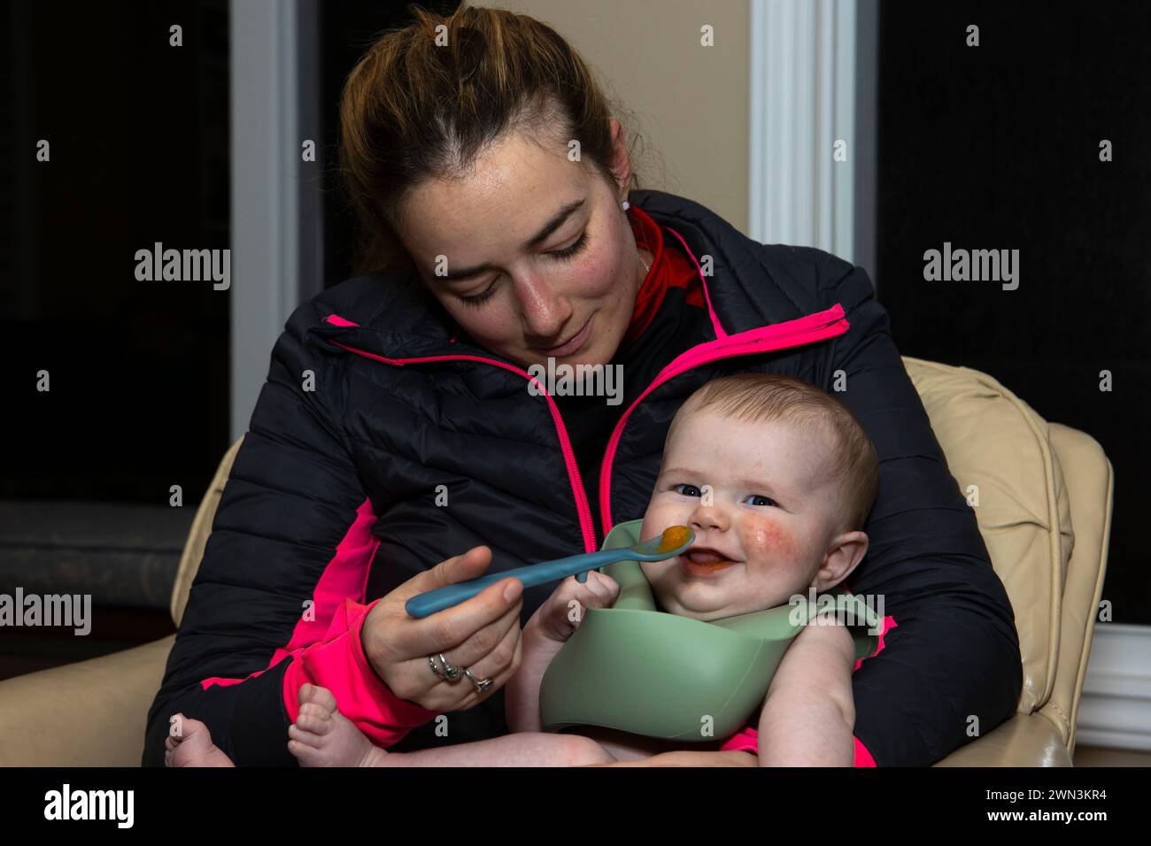 Mutter füttert einen lächelnden 4 Monate alten Jungen mit einem Löffel. Stockfoto
