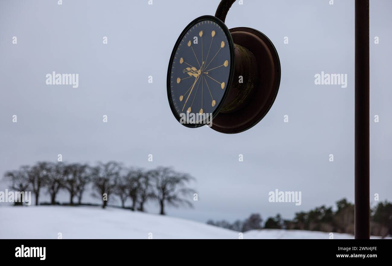 stockholmer Waldfriedhof über dem schneebedeckten Hügel hängt eine große schwarze Uhr mit goldenen Händen, die von einem Grab aus schwarzen Ulmen gekrönt ist Stockfoto