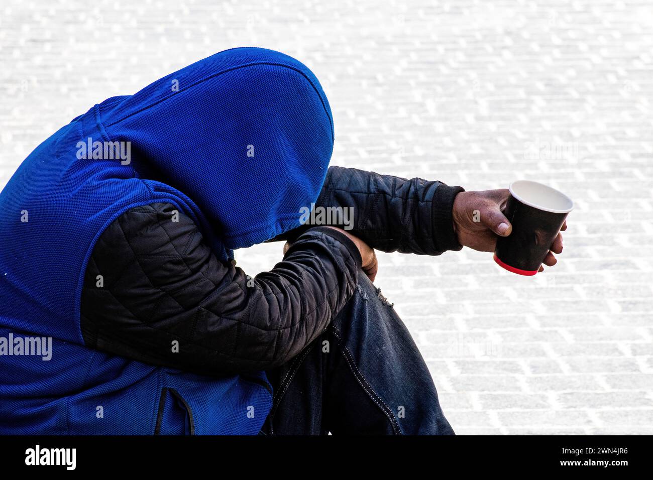 Afroamerikaner mit Jacke und königsblauem Kapuzenpulli, ausgestrecktem Arm, der eine Tasse hält und Spenden sucht. Stockfoto
