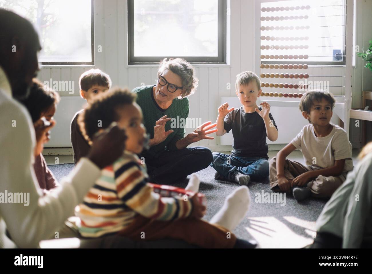 Seniorenlehrerin, die mit einer Gruppe von Vorschulkindern spielt, während sie im Kindergarten sitzt Stockfoto