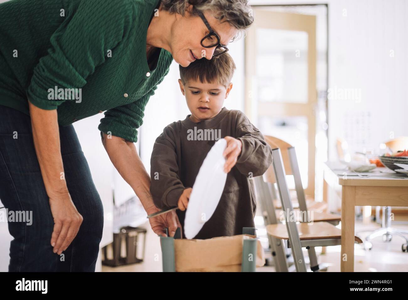 Junge, der Essensreste in den Mülleimer wirft, mit dem Lehrer im Kindergarten Stockfoto