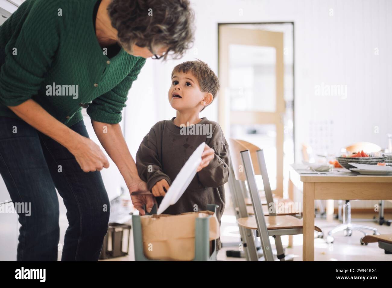 Junge, der eine Lehrerin ansieht, während er im Kindergarten Essen in die Mülltonne wirft Stockfoto