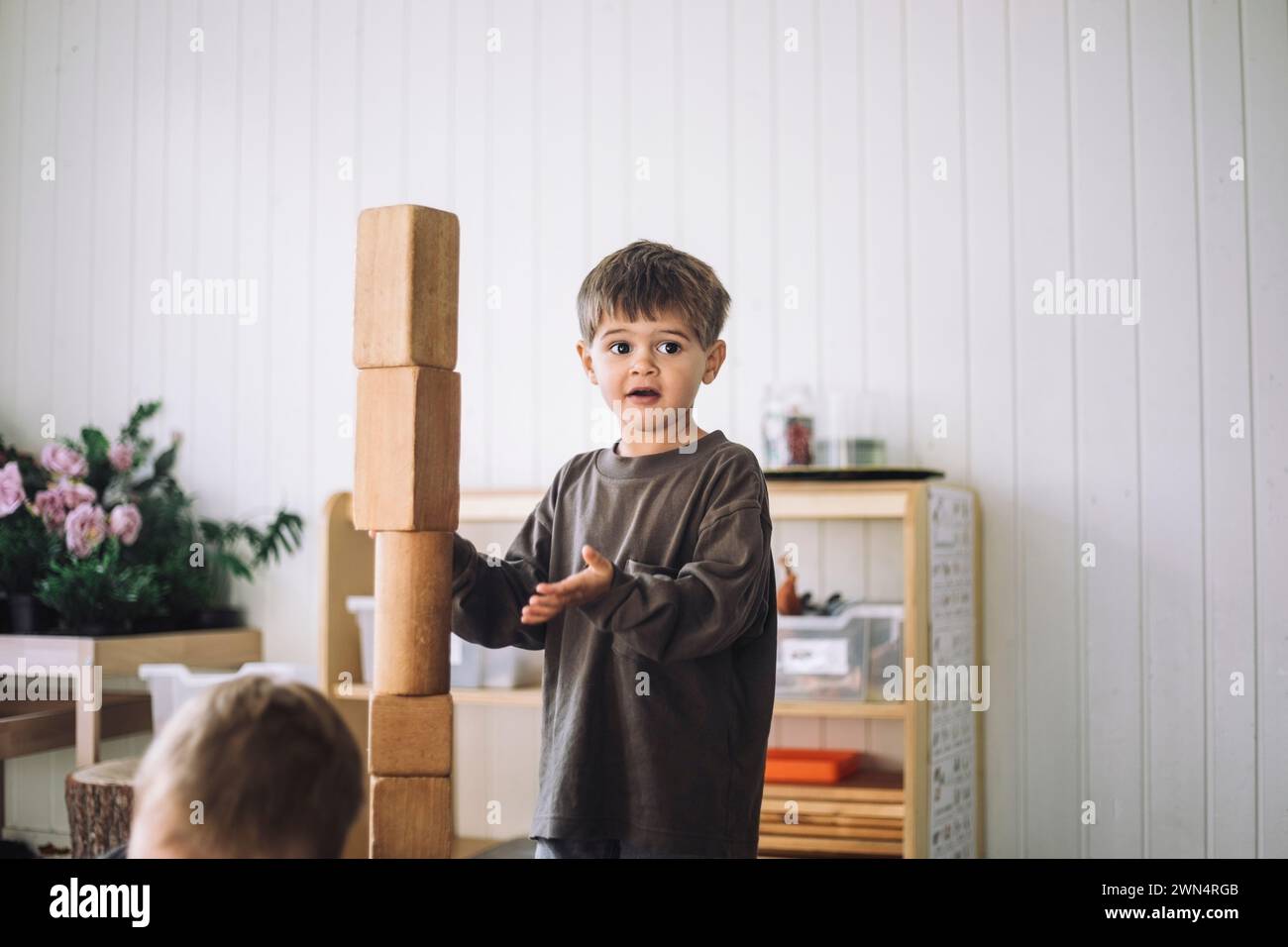 Junge, der wegschaut, während er in der Nähe eines Turms aus Spielzeugblöcken im Klassenzimmer im Kindergarten steht Stockfoto