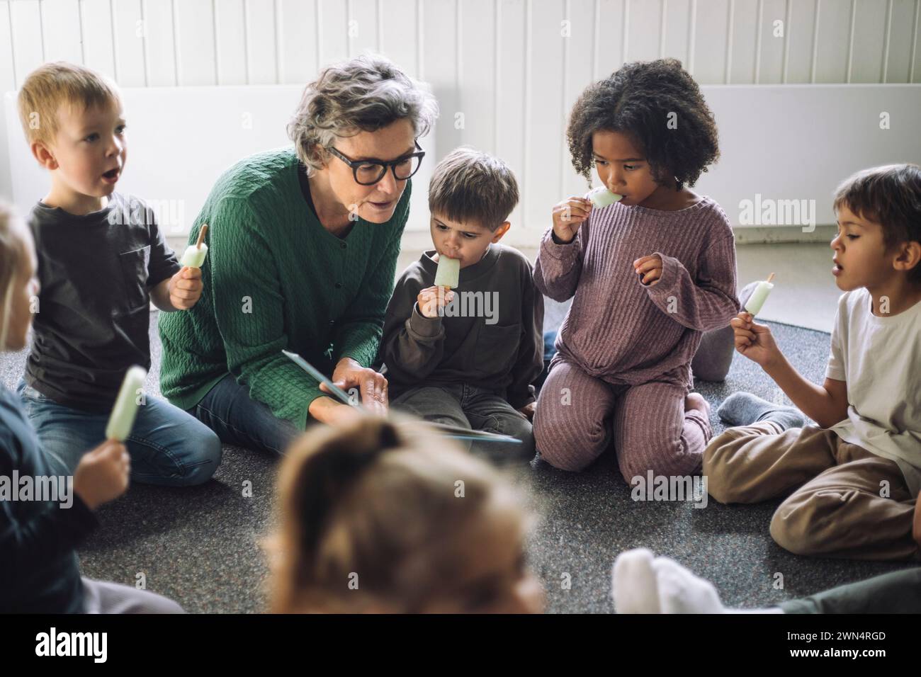 Eine Gruppe von Kindern, die Eis essen, mit einer leitenden Lehrerin, während sie im Klassenzimmer im Kindergarten saßen Stockfoto
