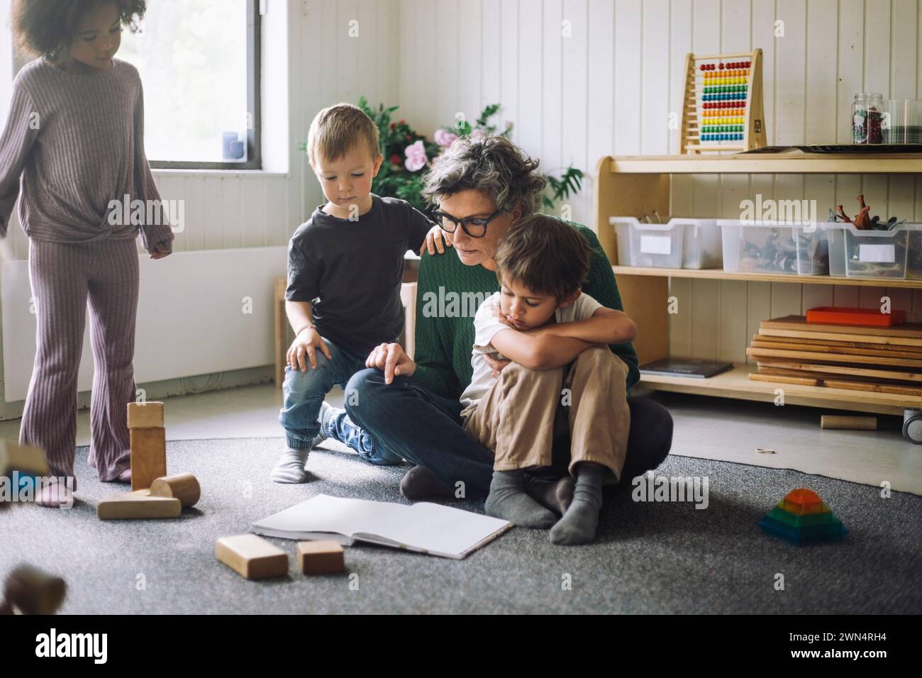 Gruppe von Kindern, die mit einer Lehrerin sitzen, die im Kindergarten Buch liest Stockfoto