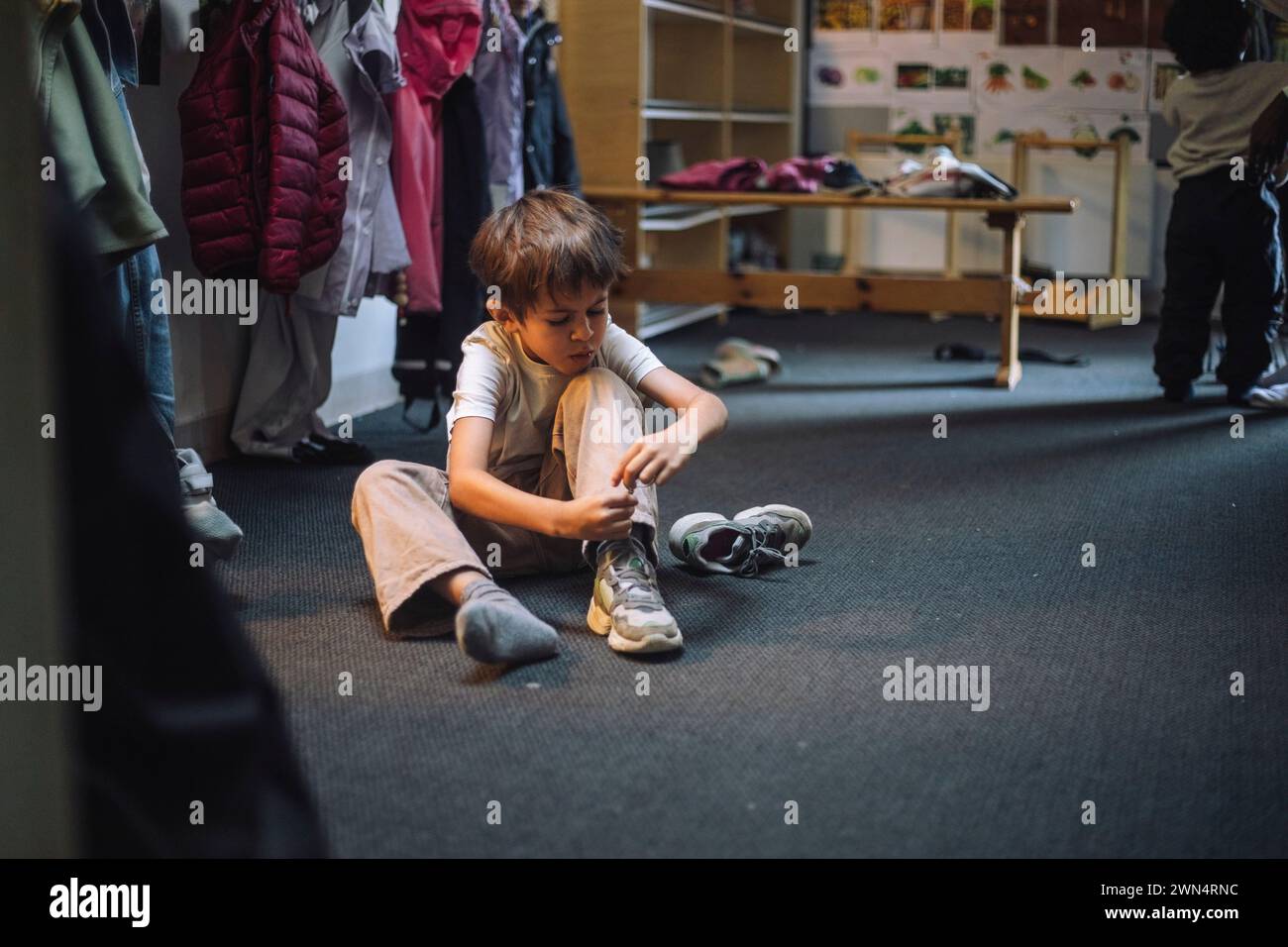 Junge, der Schnürsenkel bindet, während er auf dem Boden in der Garderobe in der Vorschule sitzt Stockfoto