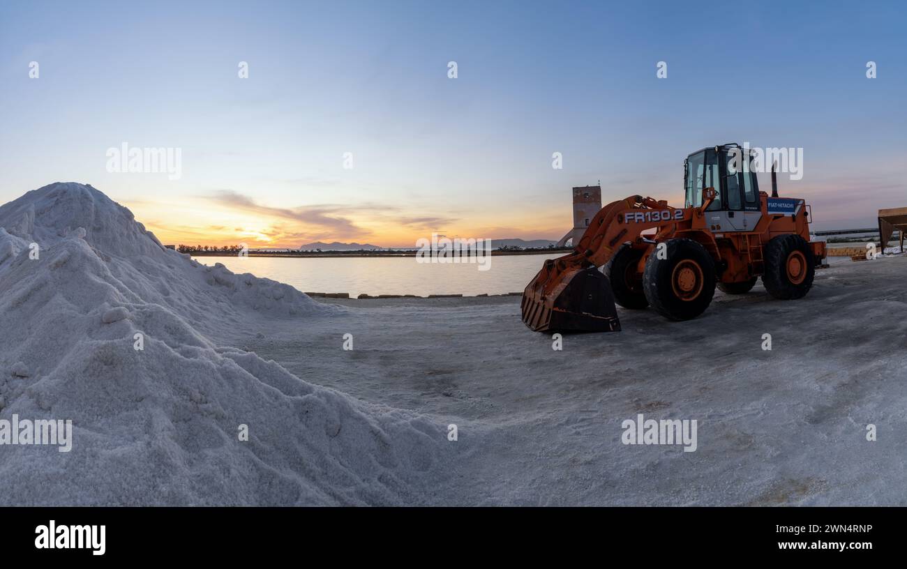 Paceco, Italien - 3. Januar 2024: Frontlader und Seesalzhaufen in der Salzebene von Trapani und Paceco bei Sonnenuntergang Stockfoto