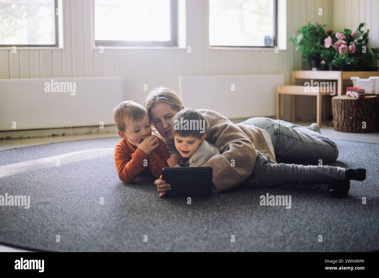 Eine Lehrerin sieht mit Jungen ein digitales Tablet, während sie sich im Vorschulalter auf dem Teppich legt Stockfoto