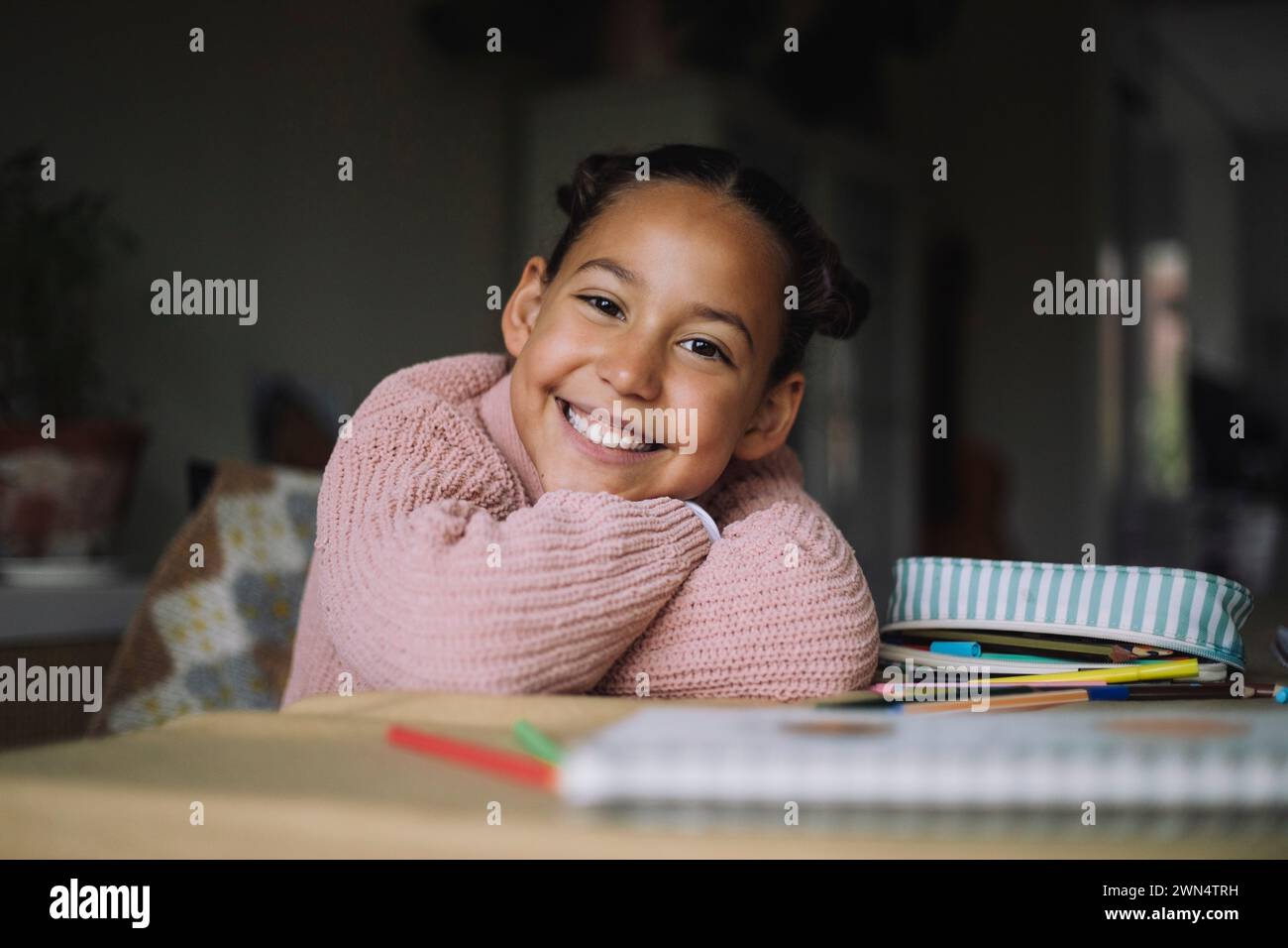 Porträt eines glücklichen Mädchens, das den Kopf auf den Armen legt, während er zu Hause mit farbigen Bleistiften auf dem Tisch sitzt Stockfoto