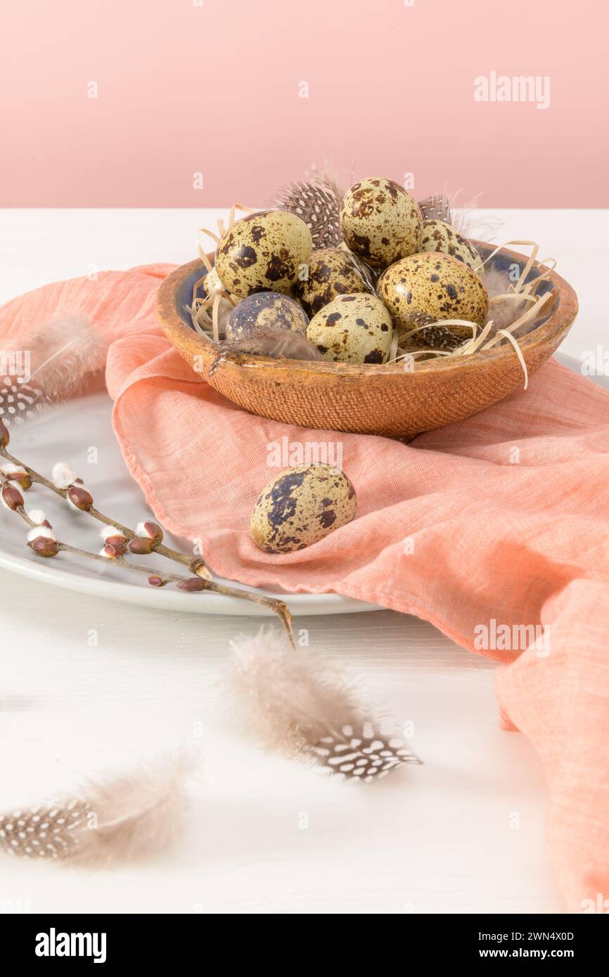 Wachteleier in einer Keramikschale mit einem pfirsichfarbenen Schal und Vogelfedern. Weißer Holztisch mit rosafarbenem Hintergrund. Hintergrund Stockfoto