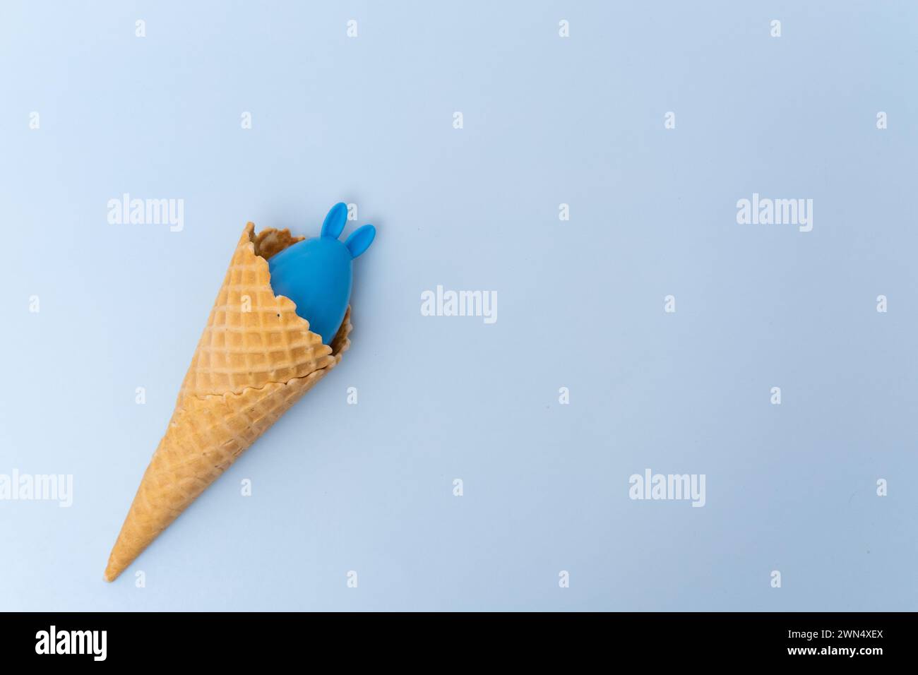 Eine Waffel-Eiskegel mit einem versteckten blauen Osterei mit Ohren wie ein Häschen auf blauem Hintergrund, Konzept, Kopierraum, flache Lay. Minimalistischer Ostergr Stockfoto