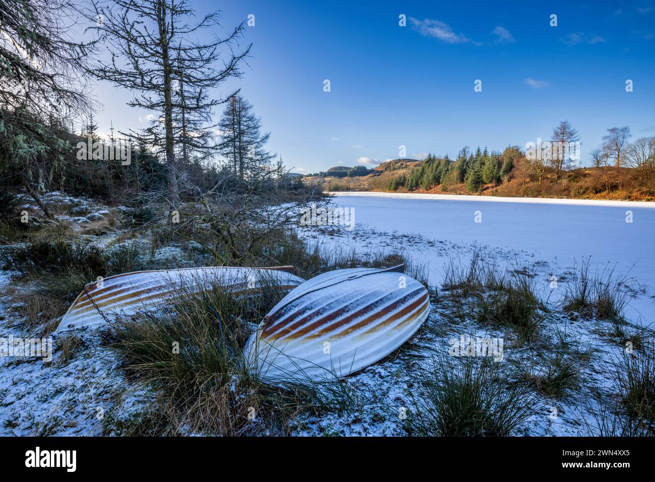 Ruderboote am Ufer eines schneebedeckten, gefrorenen Allen a’ Chip Dhuibh im Wald von Invertrossachs, Trossachs, Stirling, Schottland Stockfoto
