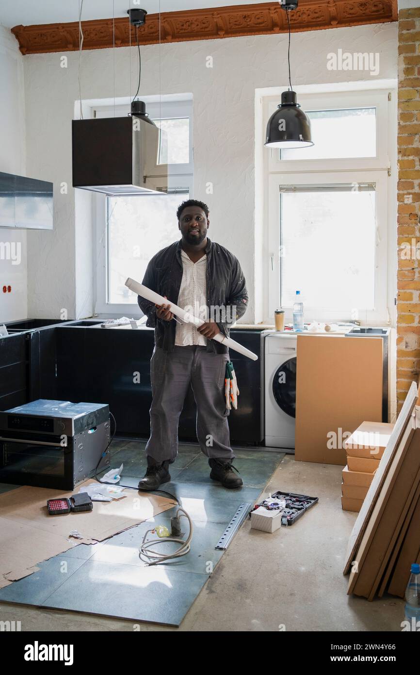 Die ganze Länge eines männlichen Zimmermanns, der einen Plan hält, der in der Küche in der Wohnung steht Stockfoto