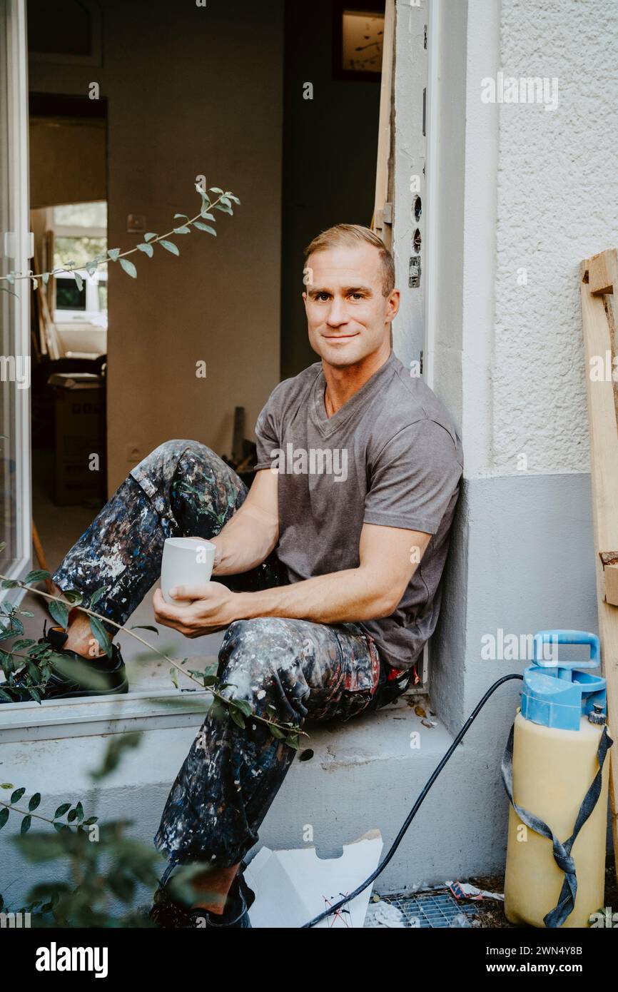 Porträt eines lächelnden männlichen Zimmermanns, der eine Kaffeetasse an der Haustür hält Stockfoto