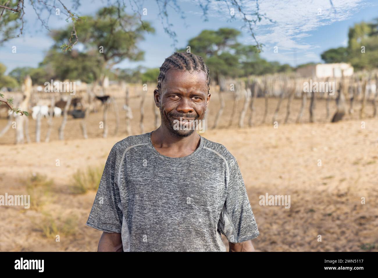afrikanischer Mann mit Zöpfen, im Dorf, vor dem Kraal mit Kleinvieh Stockfoto