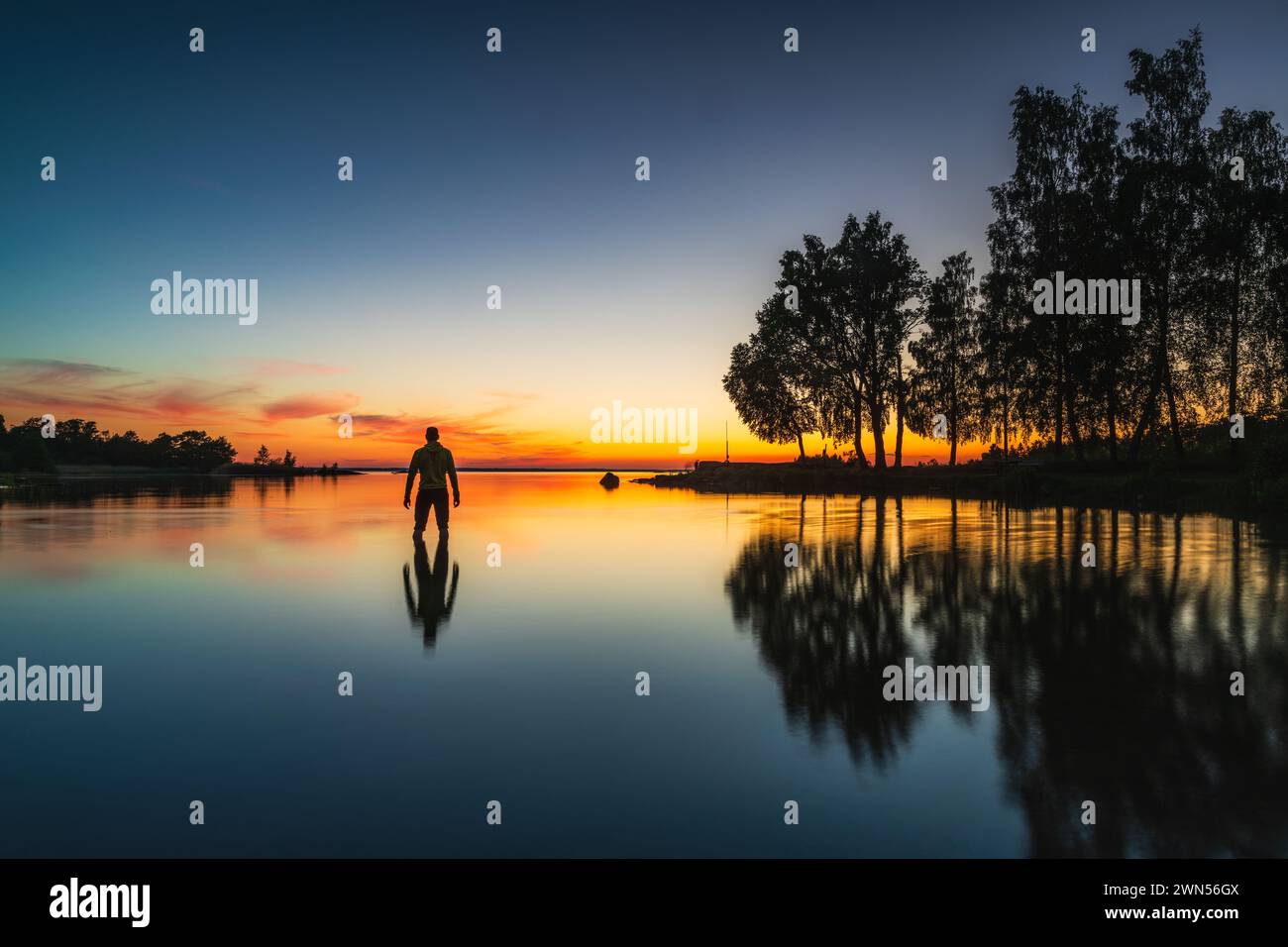 Eine ruhige Szene entfaltet sich, wenn ein Mann am Rand eines ruhigen Sees steht, und die markanten Farben des Sonnenuntergangs spiegeln sich auf der Wasseroberfläche wider. Die Stockfoto