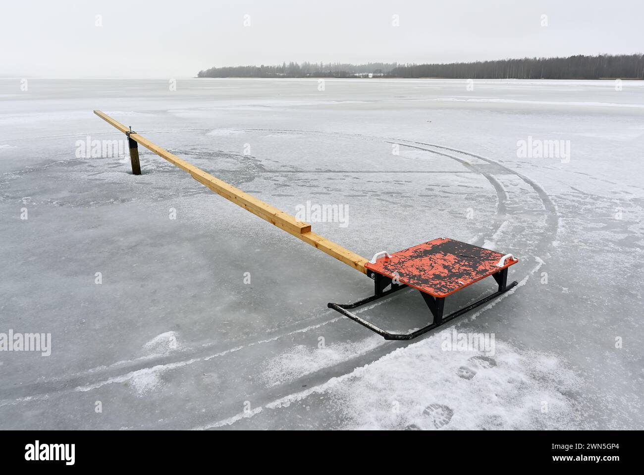 Ein einsamer gefrorener See mit einem einsamen roten Schlitten, der an einer langen Holzplanke befestigt ist, schafft einen starken Kontrast Stockfoto