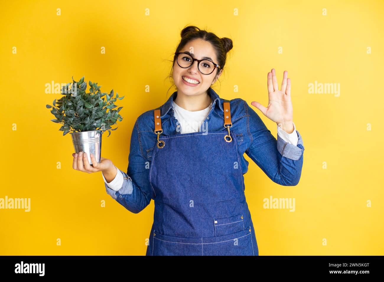Junge kaukasische Gärtnerin, die eine Pflanze isoliert auf gelbem Hintergrund hält und Star-Trek-Freak-Symbol macht Stockfoto