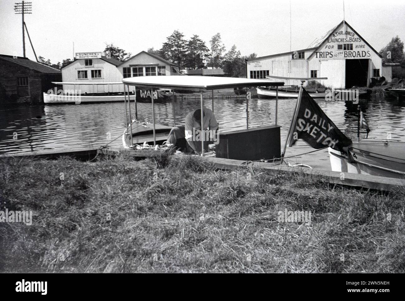 1950er Jahre, historisch, Bootswerften auf den Norfolk Broads... auf der rechten Seite sehen Sie die Werft von A Thrower, die Starts und Boote zum Verleih und Trips zu den Broads bietet. Im Vordergrund ein Mann auf einem kleinen Boot, mit der Flagge Daily Sketch, eine nationale Boulevardzeitung der damaligen Zeit. Stockfoto