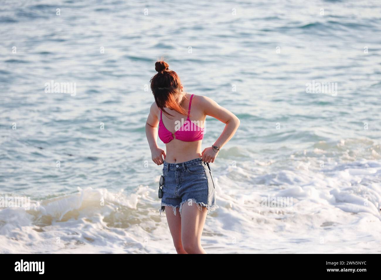 Asiatisches Mädchen in Jeans-Shorts und rosa Bikini, die am Strand auf Surfwellen-Hintergrund stehen Stockfoto