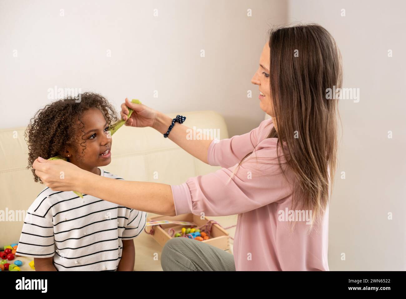 Eine pflegende Szene entfaltet sich, während eine Mutter sorgfältig die lockigen Haare ihrer afroamerikanischen Tochter stylt, deren Bindung in ihren fröhlichen Ausdrucksformen deutlich wird. Hg Stockfoto