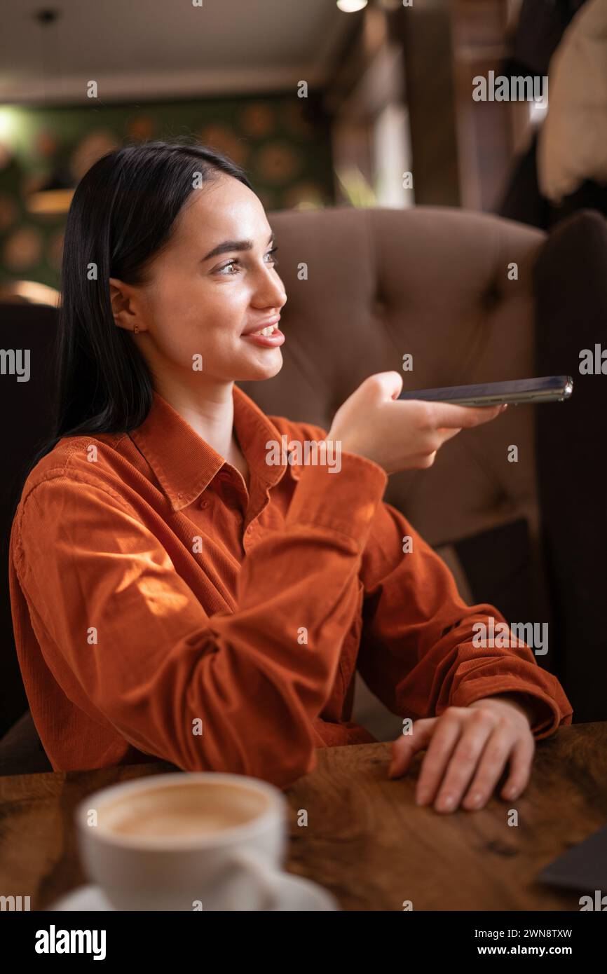 Mädchen, das online eine Smartphone-Spracherkennung nutzt und im Café sitzt und an einem Laptop arbeitet. Sprachnachricht. Stockfoto