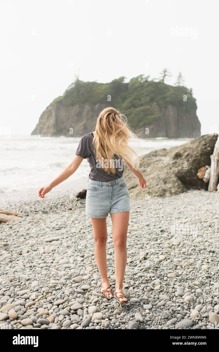 Junge Frau, die an einem felsigen Strand läuft, mit langen Haaren, die im Wind wehen Stockfoto