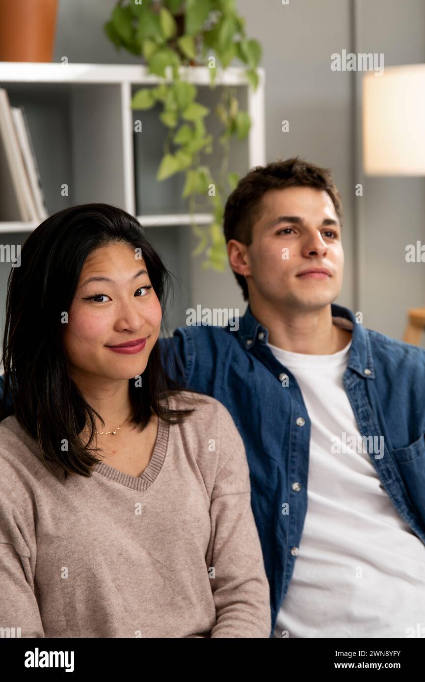 Lächelnder junger Erwachsener vielfältiges Paar, das sich zu Hause auf dem Sofa entspannt. Frau, die in die Kamera schaut, neben einem glücklichen Mann, der auf einer Couch sitzt Stockfoto
