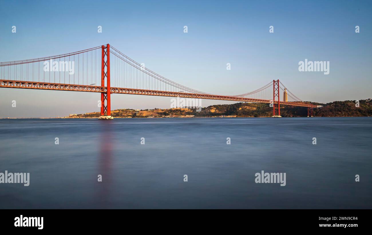 Ruhiger Blick auf eine rote Hängebrücke bei Sonnenuntergang über ruhigem blauem Wasser mit Reflexionen, Lissabon, Brücke 25 de Abril Stockfoto