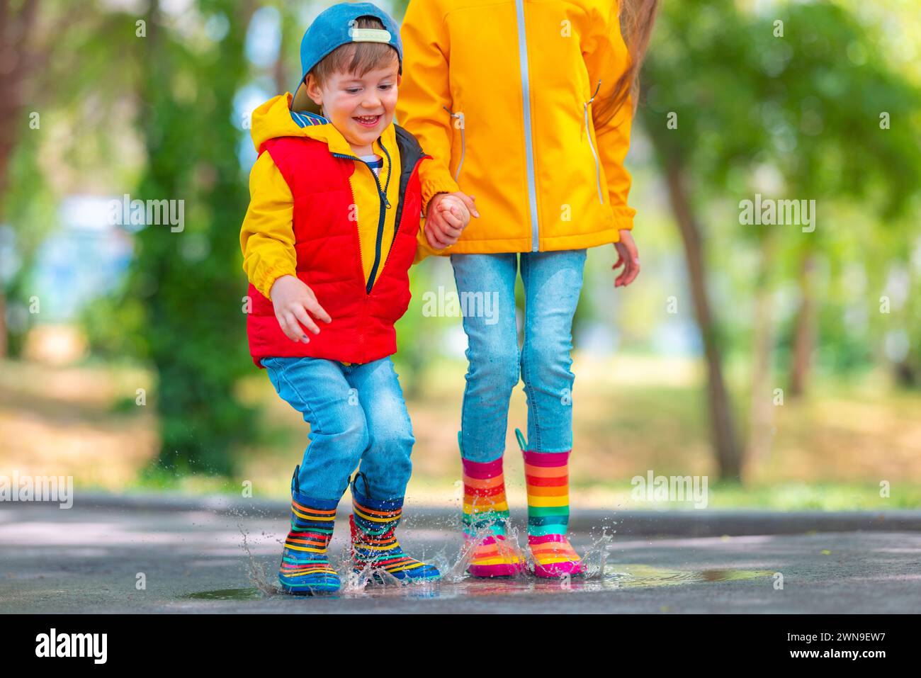 Glückliche Kinder Mädchen und Junge mit Regenschirm und farbigen Gummistiefeln spielen draußen und springen in schmutzigen regnerischen Pfützen Stockfoto