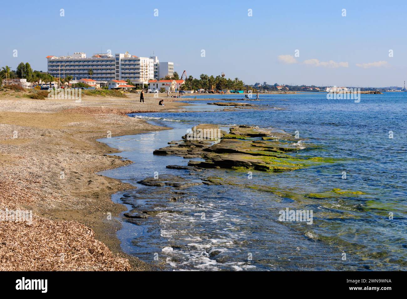 Voroklini Strand mit Blick auf das Lordos Beach Hotel. Felsen zeigen sich im Meer, Leute laufen am Strand. Larnaka, Zypern. Februar 2024 Stockfoto