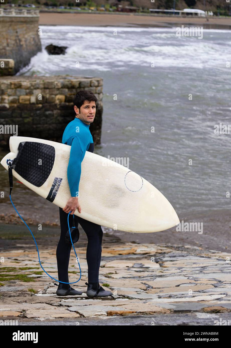 Ein Surfer im Neoprenanzug trägt sein Surfbrett unter den Armen und blickt mit Vorfreude auf die Meereswellen zurück, bereit zum Surfen. Stockfoto