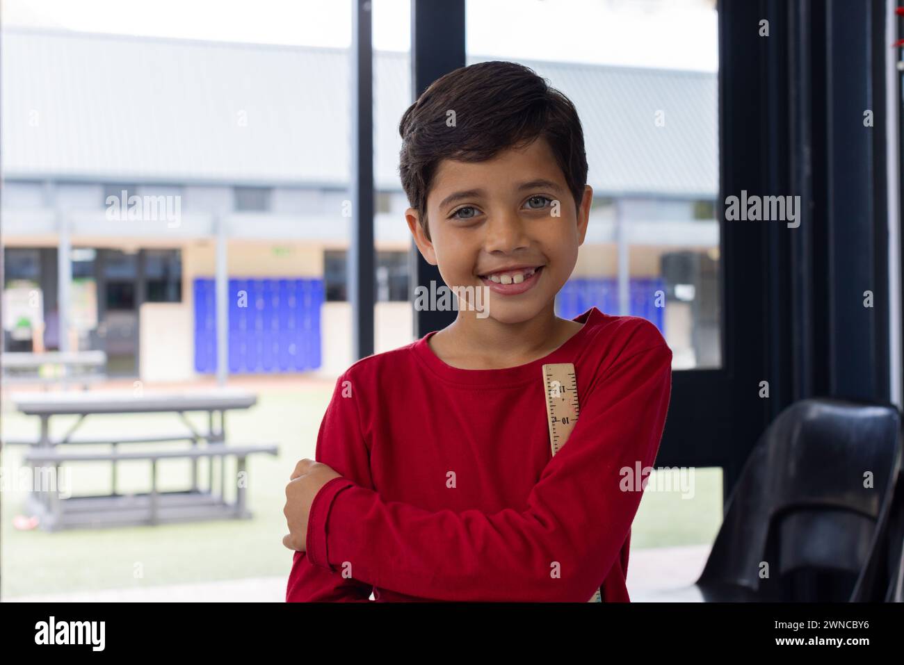 Ein birassischer Junge mit einem hellen Lächeln, der ein rotes Hemd trägt, steht selbstbewusst in der Schule Stockfoto