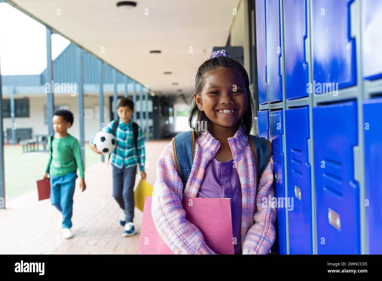 Ein birassisches Mädchen mit einem lila Rucksack lächelt an den blauen Schließfächern der Schule, Jungs im Hintergrund Stockfoto