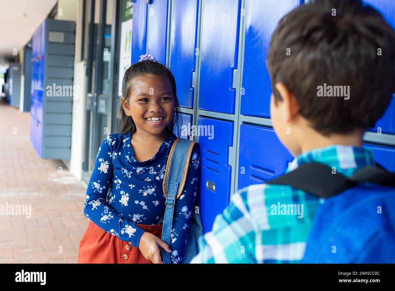 Ein birassisches Mädchen mit einem hellen Lächeln spricht mit einem birassischen Jungen an blauen Schließfächern Stockfoto
