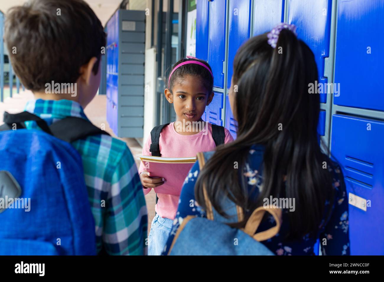 Ein birassisches Mädchen mit rosa Stirnband spricht mit ihren Kollegen an blauen Schließfächern und hält ein Notizbuch Stockfoto