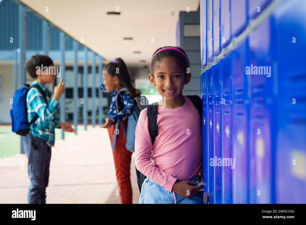 Ein birassisches Mädchen mit einem rosa Rucksack lächelt neben den blauen Schließfächern der Schule, ihr dunkles Haar ist zurückgebunden Stockfoto