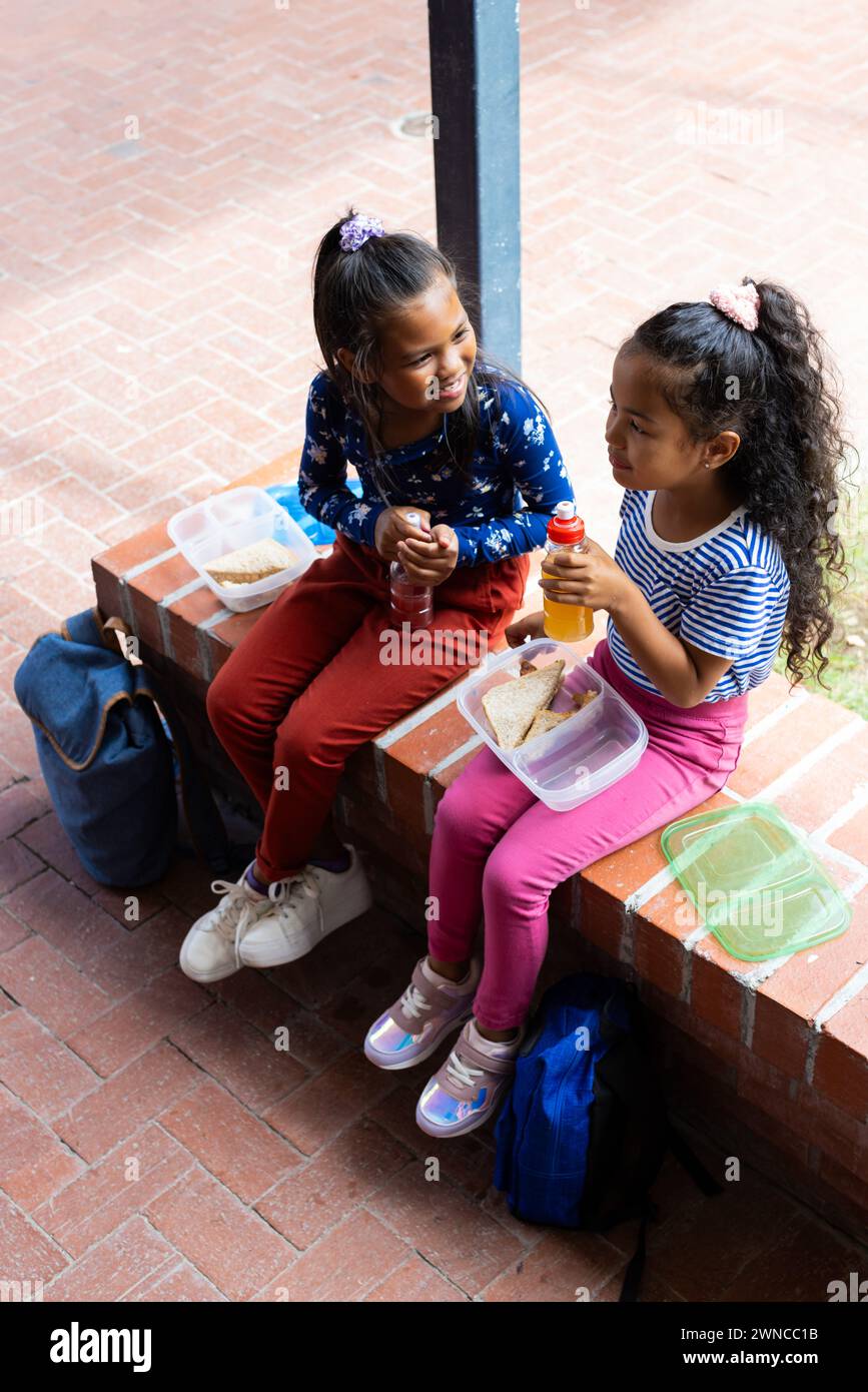 Zwei rassistische Mädchen genießen eine Mittagspause im Freien in der Schule und teilen sich Lächeln und Sandwiches Stockfoto