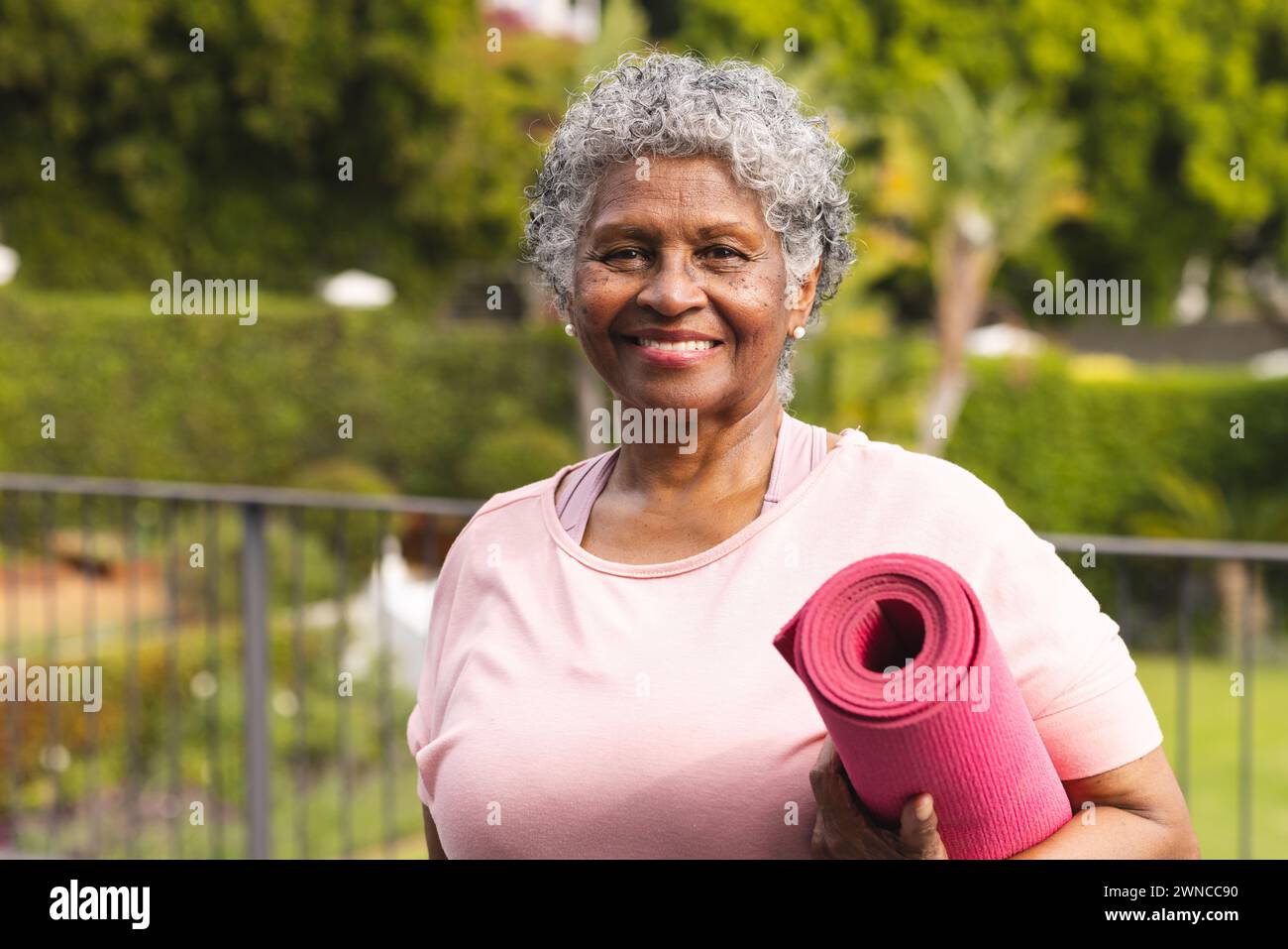 Ältere birassische Frau mit grauen Haaren hält draußen eine rosafarbene Yogamatte Stockfoto