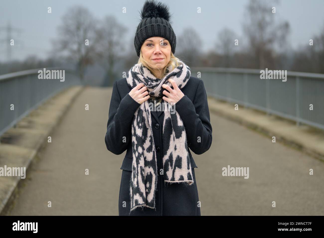 Die blonde Dame trägt eine schwarze Jacke und einen schwarzen Wackelhut, der ihren Schal hält, während sie auf einer Brücke steht Stockfoto