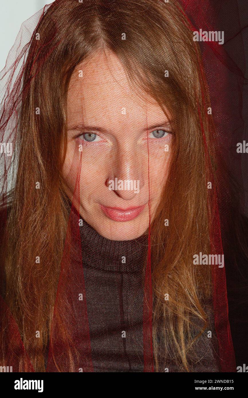 Vertikale Zusammensetzung. Porträt einer erwachsenen jungen Frau mit langen blonden Haaren und grauen Augen und einem roten, transparenten Tuch auf dem Kopf. Stockfoto