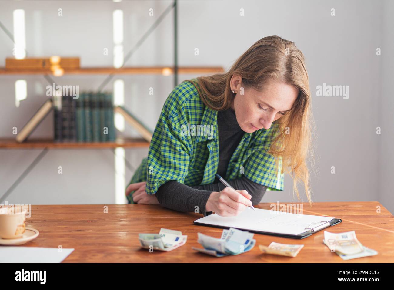 Eine junge hübsche Dame macht nachdenklich und aufmerksam Notizen vor dem Hintergrund von gestaltetem und gezähltem Geld. Stockfoto